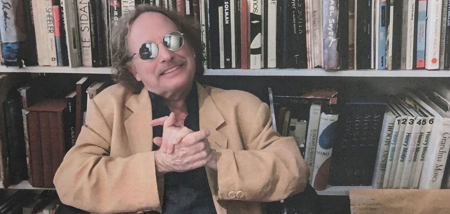 Jeffrey Bergen seated in front of a wall of art books, hands clasped, smiling.