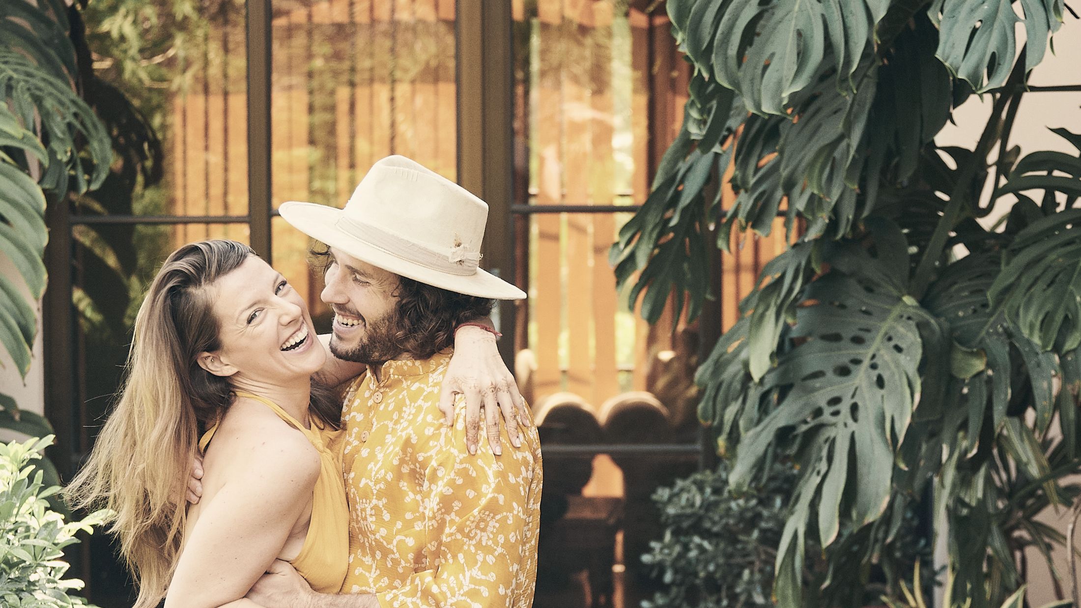 Kayla and Vaughn laughing together in a plant-filled courtyard, both dressed in yellow.